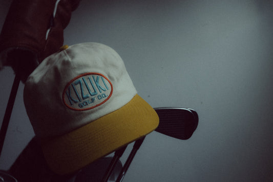 A white and yellow baseball cap with a circular 'Kizuki Golf Co.' logo patch, resting on a golf club against a dark gray background.
